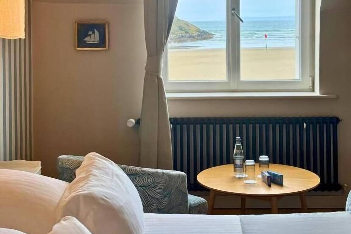 A hotel room with a bed, a round table with a bottle and glasses, a chair, a view of the beach, and a radiator.