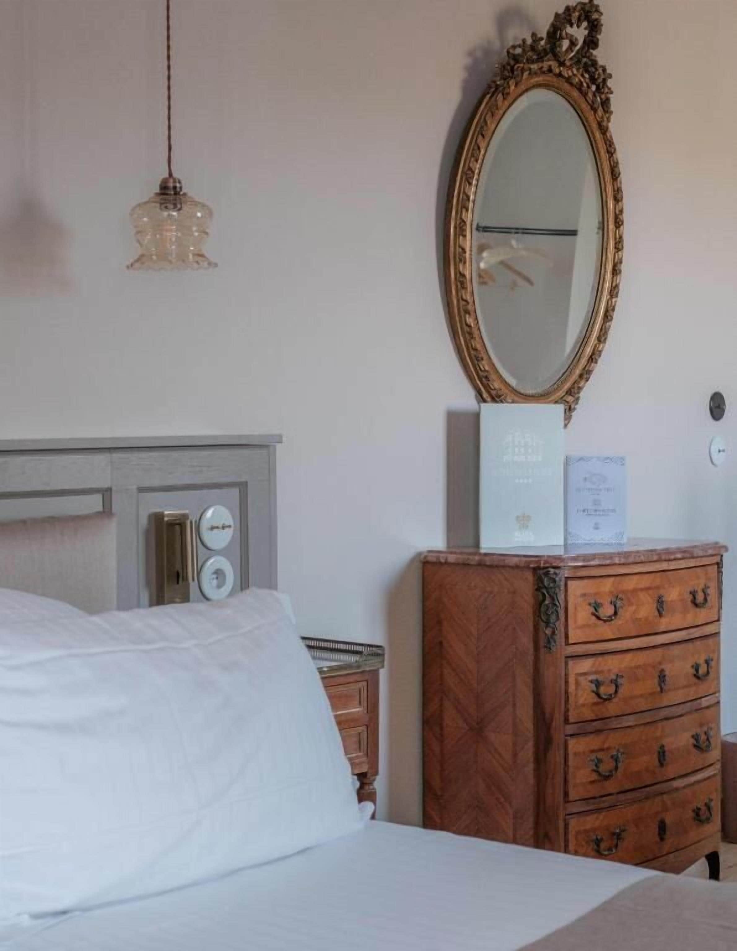 Habitación de hotel con cama, cómoda de madera con cajones, espejo y un reloj de pared.