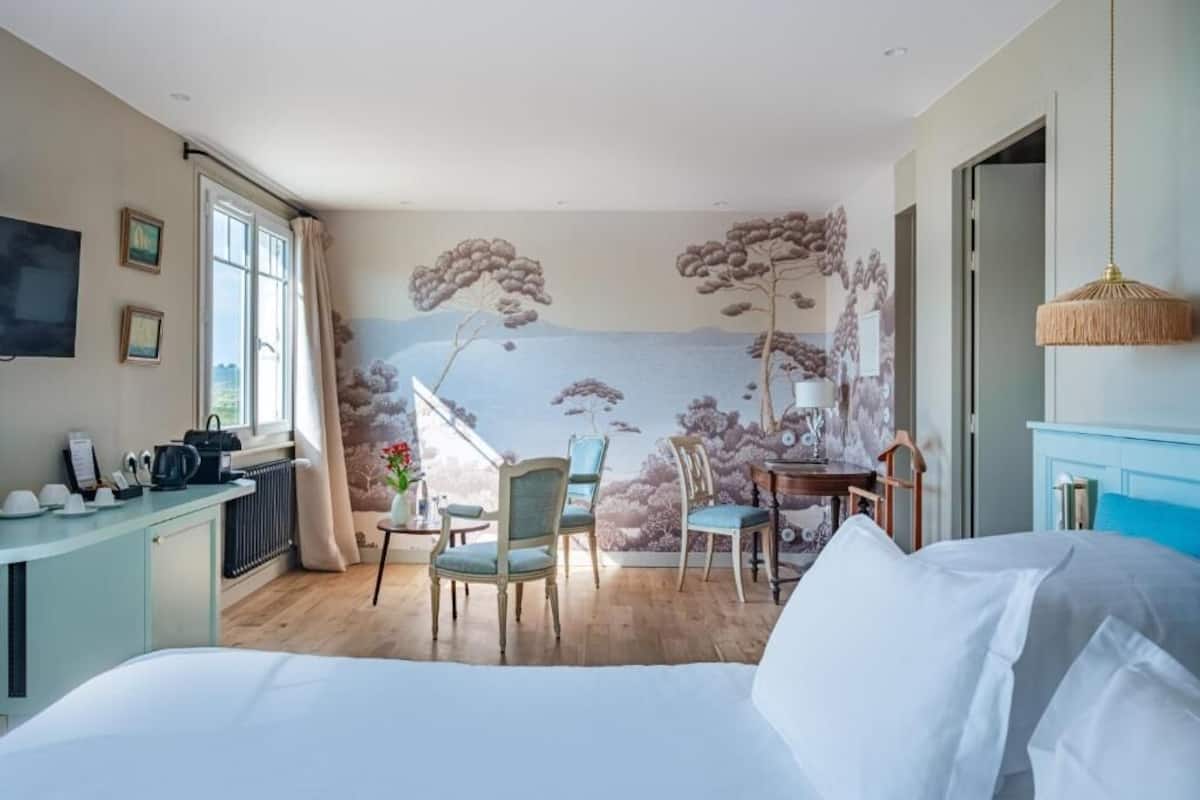 A bedroom with a bed, a desk, chairs, a view of the outdoors, and a mural on the wall.