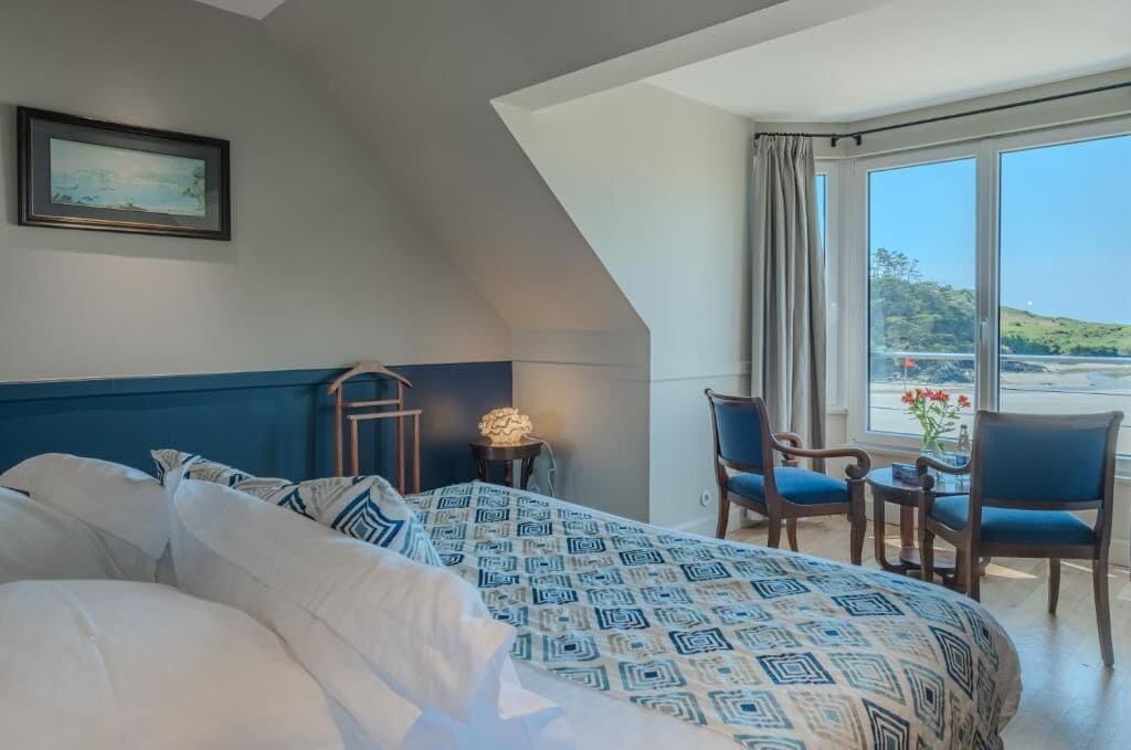 A bedroom with a bed, a desk, chairs, a lamp, and a window with a view.