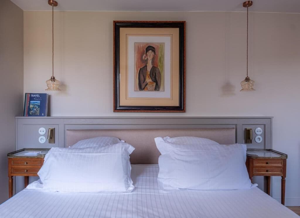 Habitación de hotel con cama, dos mesitas de noche, un cuadro en la pared y un libro sobre una de las mesitas.