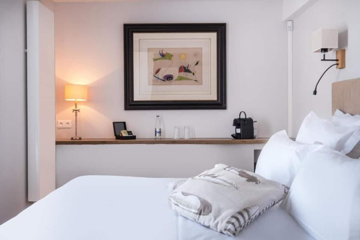 A hotel room with a bed, a bedside table with a lamp, a framed artwork, and a coffee machine.