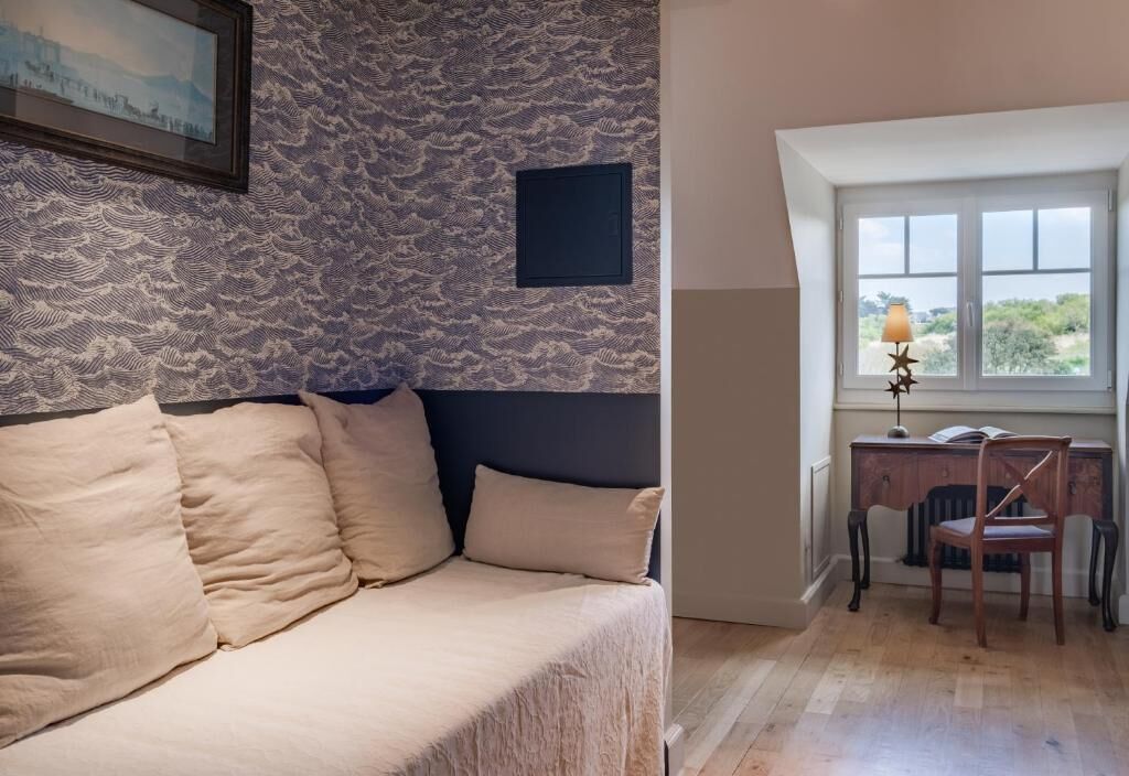 Una habitación con pared estampada, una cama con ropa de cama color beige, un escritorio con una silla y una ventana con vistas.