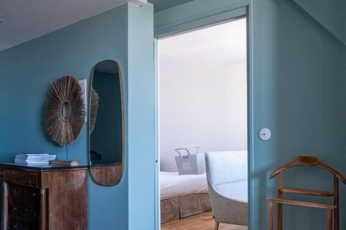 A room with a wooden floor, a desk with a mirror, a bed with a grey headboard, and a wooden ladder.