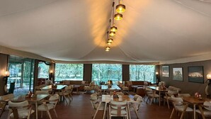 A spacious indoor dining area with multiple tables and chairs, large windows, and hanging pendant lights. - Serenity Mara Legends Camp (Maasai Mara)