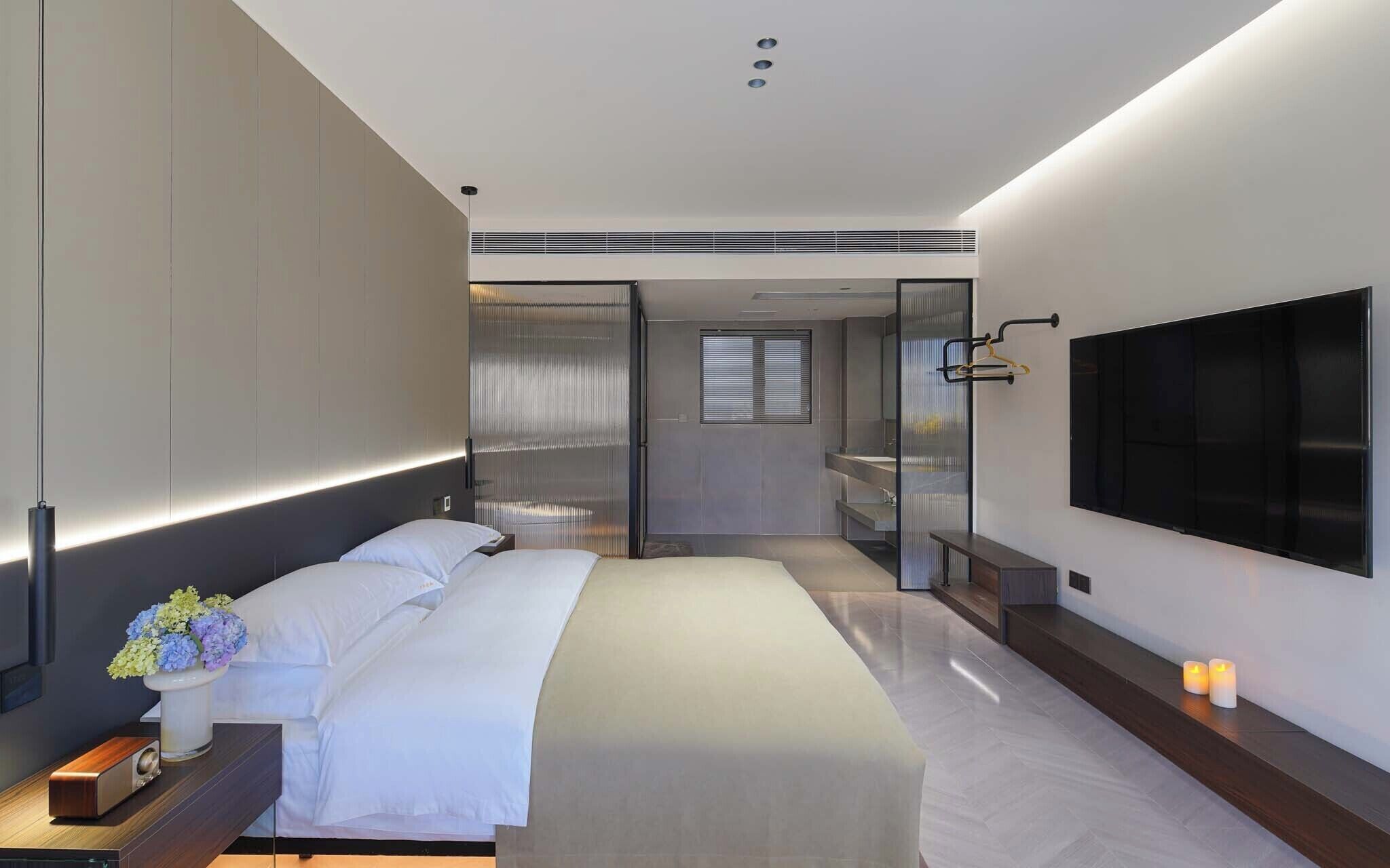Quarto moderno com cama, mesa de cabeceira, televisão e porta de vidro deslizante para um banheiro.