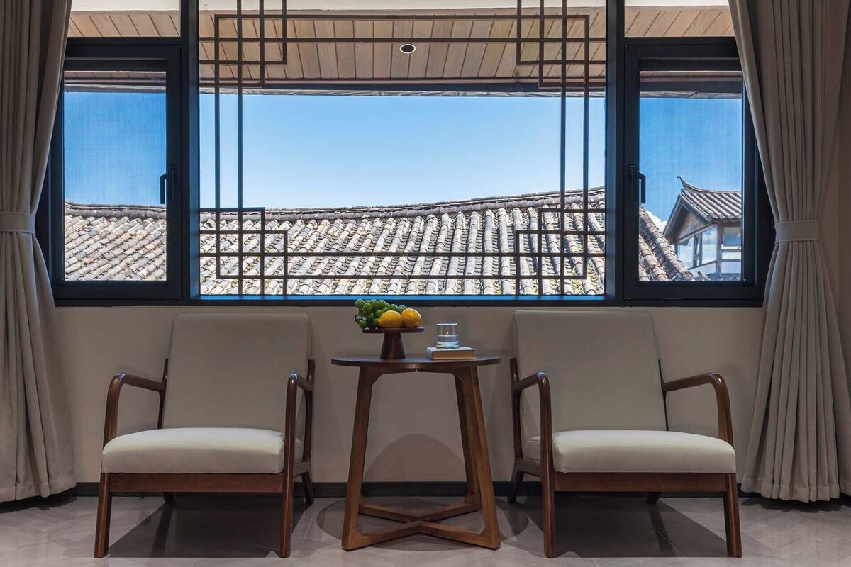 Una habitación con dos sillas, una mesa pequeña con fruteros y vistas a tejados tradicionales con tejas a través de amplias ventanas.