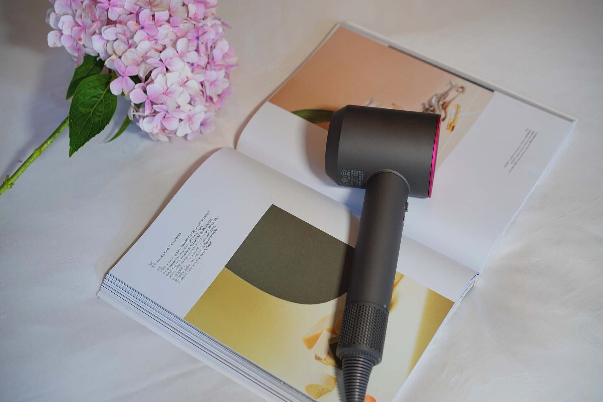 A book with a black object on top, possibly a hairdryer, next to a pink flower.