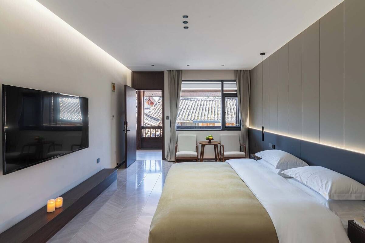 Una habitación de hotel moderna con una cama grande, un televisor de pantalla plana, una pequeña mesa con sillas y vistas a la arquitectura tradicional fuera de la ventana.