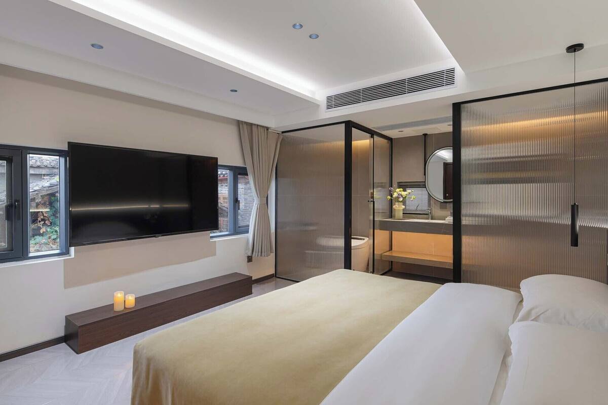 Una habitación de hotel moderna con una cama grande, un televisor de pantalla plana, un baño con espejo y un ventanal con cortinas.