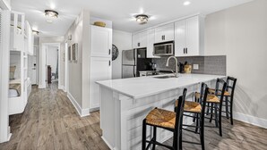 A modern kitchen with white cabinetry, a large island with bar stools, a microwave, and a refrigerator. - Barefoot Bliss | Lakefront Villa Steps from Miramar Beach (Miramar Beach)