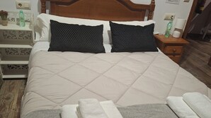 Una cama bien hecha con cabecera de madera, dos almohadas, una mesita de noche con lámpara y un pequeño mueble con una botella de agua.