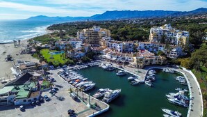 A coastal marina with boats, a beach, and buildings with balconies overlooking the water.