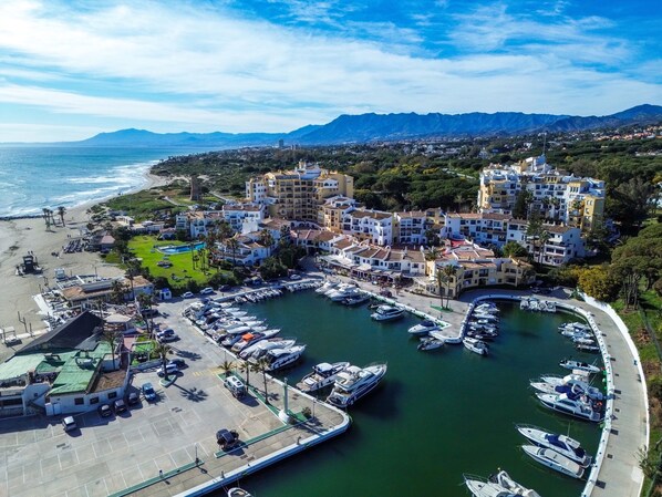 A coastal marina with boats, a beach, and buildings with balconies overlooking the water.