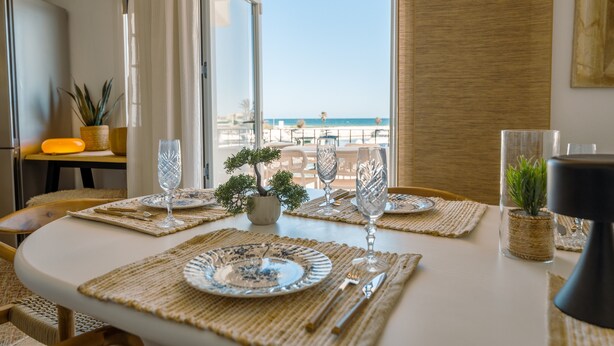 A dining table set with plates, cutlery, and wine glasses, with a view of the beach and ocean through a glass door.