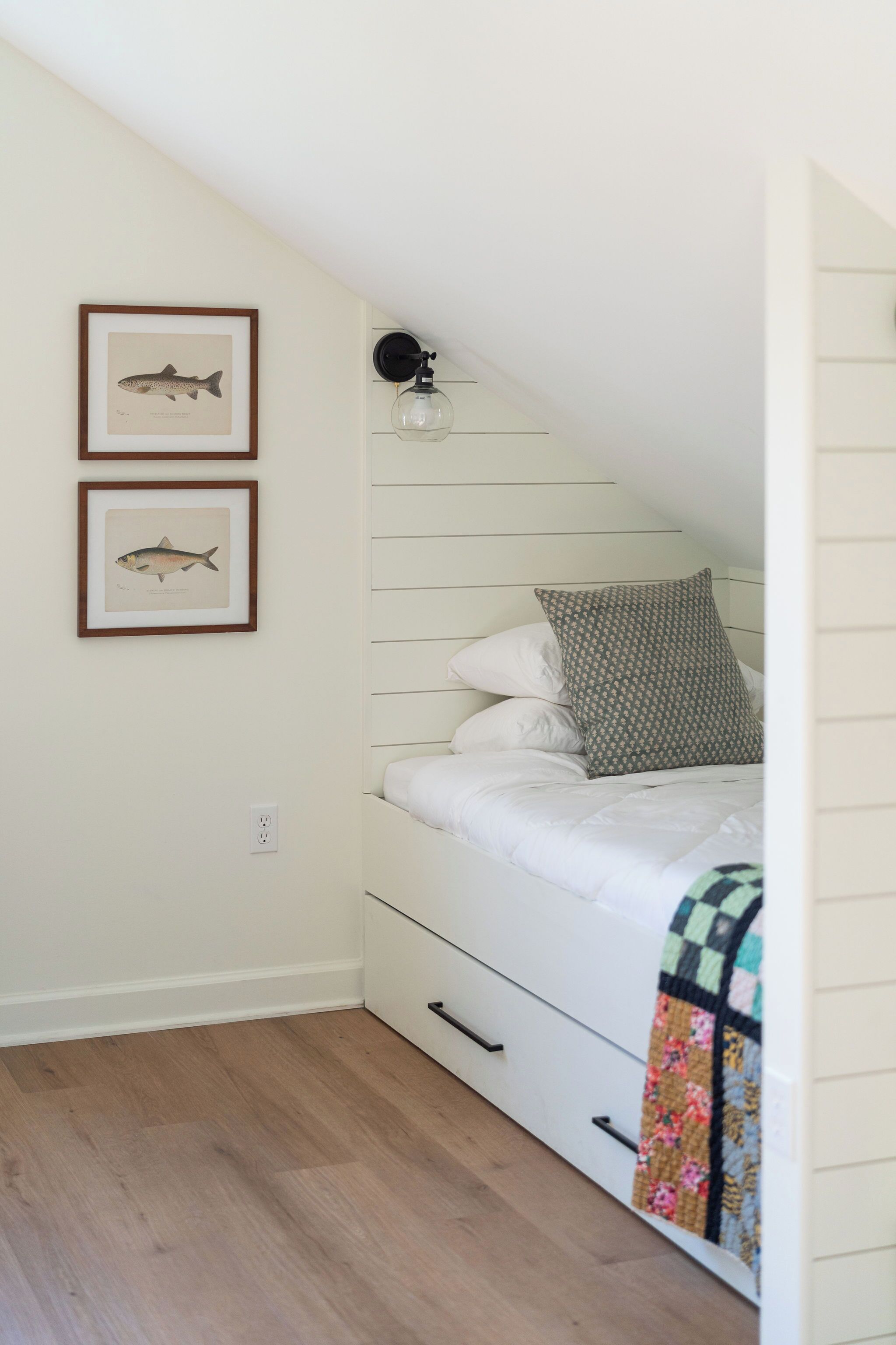 Ein Schlafzimmer mit schrägen Wänden, einem Bett mit weißer Bettwäsche, zwei gerahmten Fischbildern an der Wand, einer Nachttischlampe und einer Steppdecke auf dem Bett.
