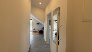 Un pasillo con una lámpara en el techo, una puerta con espejo y vista a una habitación con sofá y lámpara.