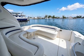 A boat interior with a cushioned seating area, a small table, and a view of the marina with other boats and a distant shoreline. - Family Yacht Stay · Sleeps 6 · Pool, Gym & Beach (Key West)