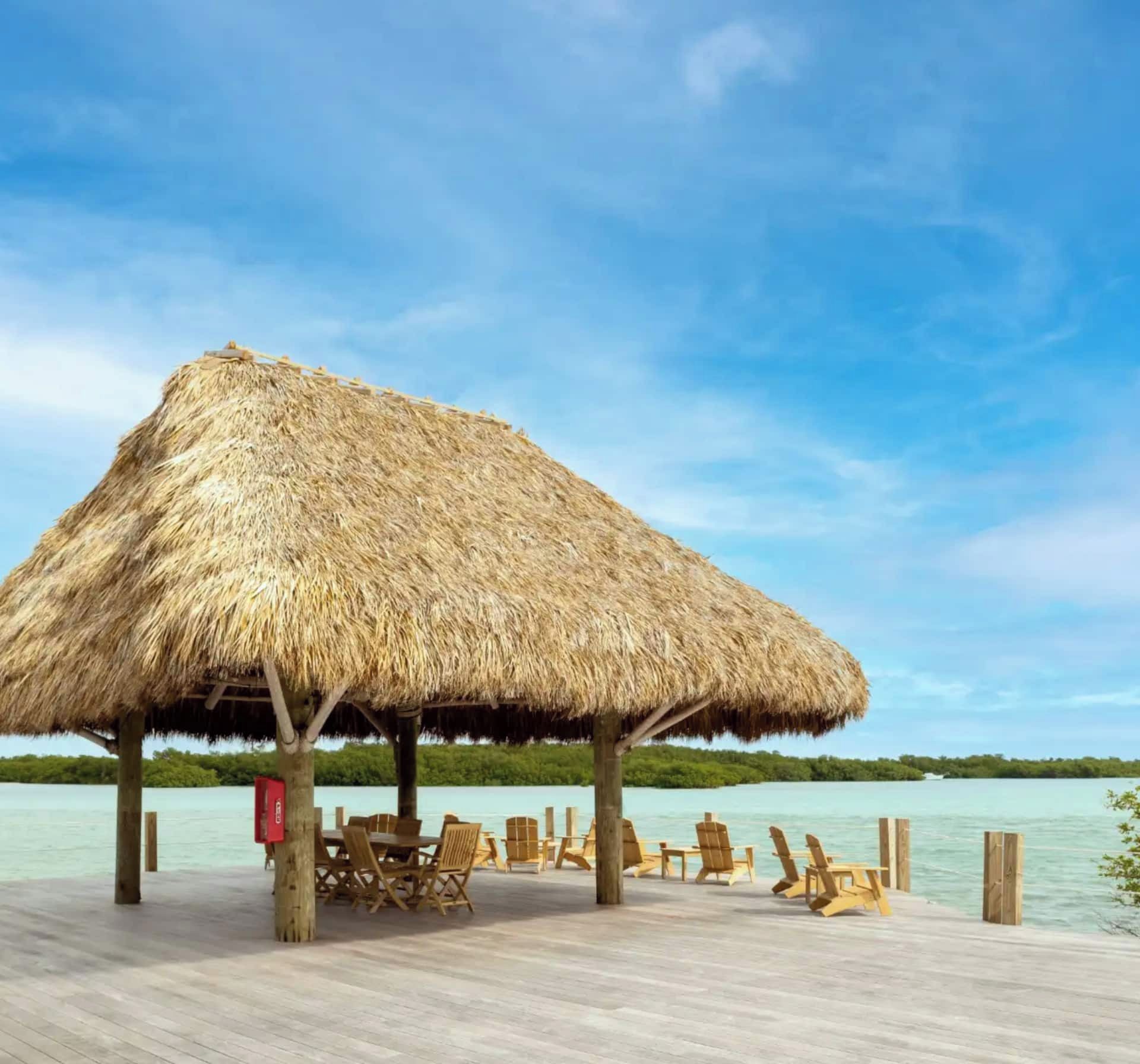 A thatched-roof structure with wooden chairs and tables on a wooden deck by a body of water.