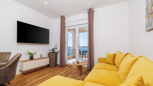 A modern living room with a yellow sofa, a flat-screen TV, a wooden floor, and a balcony with a view. - Apartment in Dramalj with balcony, sea view and pool (Crikvenica)