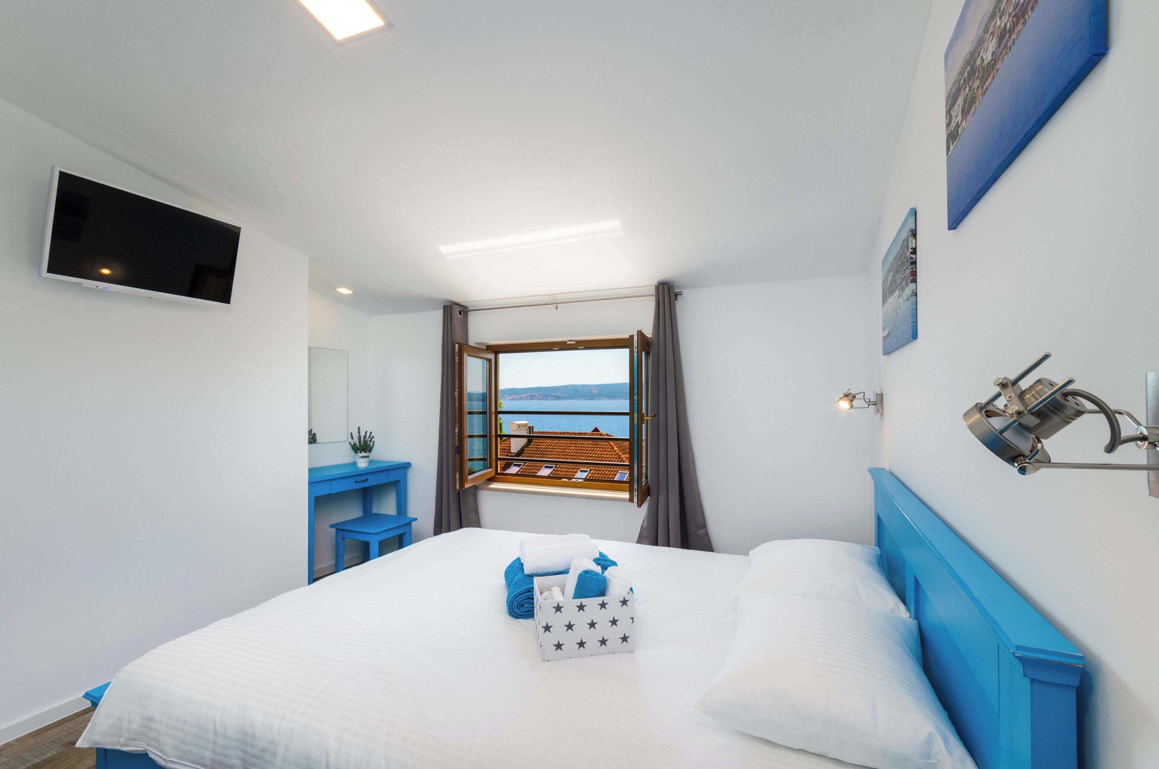 A bedroom with a bed, a desk, a chair, a TV, a window with a view, and a wall-mounted lamp.