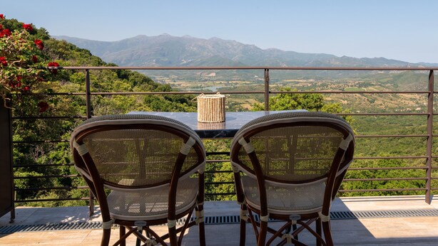Un balcon avec deux fauteuils en osier et une table, donnant sur un paysage montagneux.