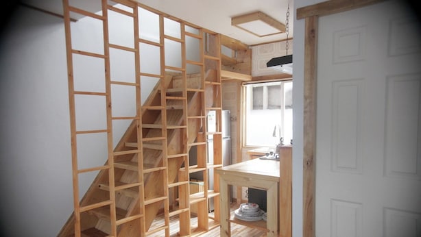 Una escalera de madera con barandilla de rejilla, una zona de cocina con lavabo y nevera, y una puerta blanca.