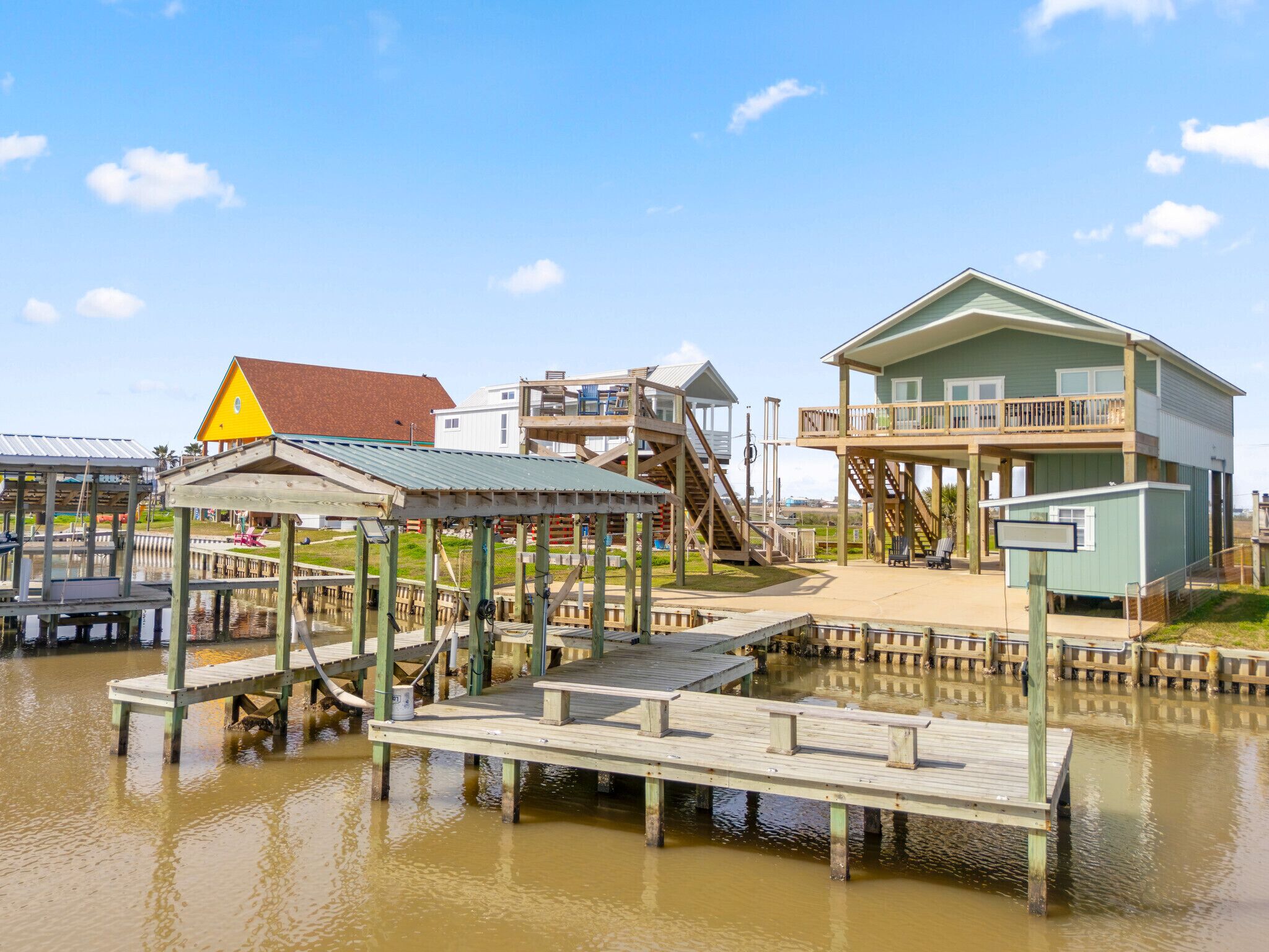 A waterfront property with a wooden dock, a multi-level house with a balcony, and a small building with a triangular roof.