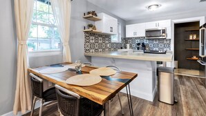 A modern kitchen with a dining area, featuring a wooden table, wicker chairs, a built-in microwave, and a trash bin. - 3 BR Tampa Pool Home - Putting Green |Firepit |Coffee Bar (Tampa)