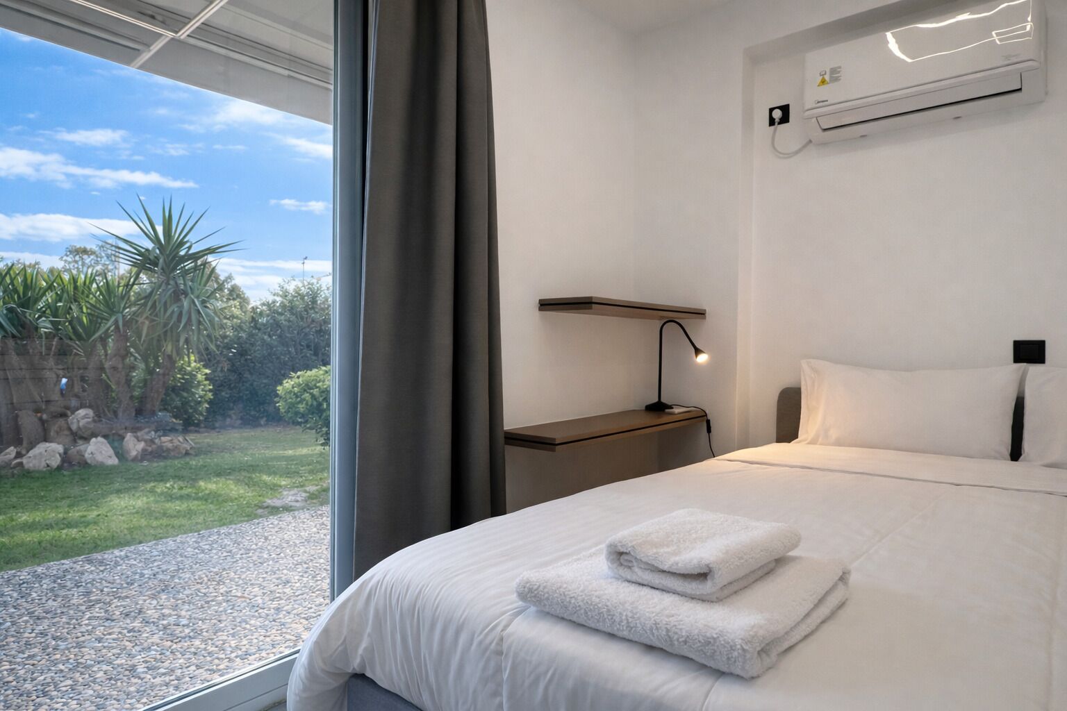A modern bedroom with a large window, a bedside table with a lamp, and a neatly made bed with towels.