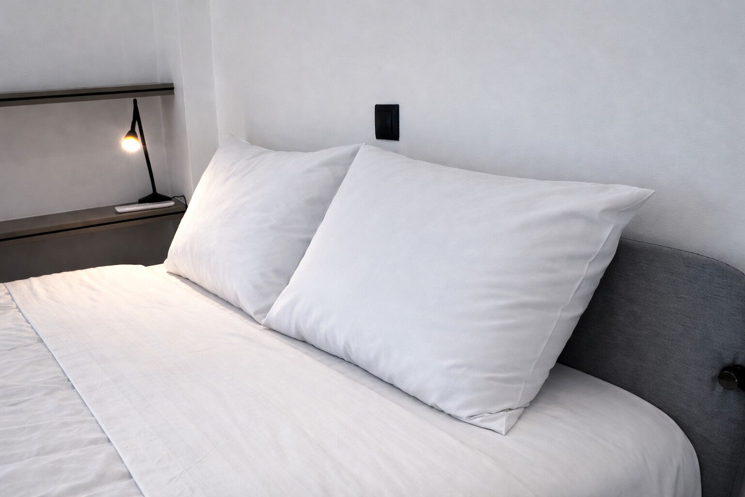 A neatly made bed with white bedding and pillows, a bedside lamp, and a modern wall-mounted switch.