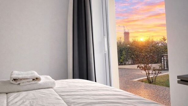 A hotel room with a bed, a window with curtains, and a view of a sunset over a cityscape.