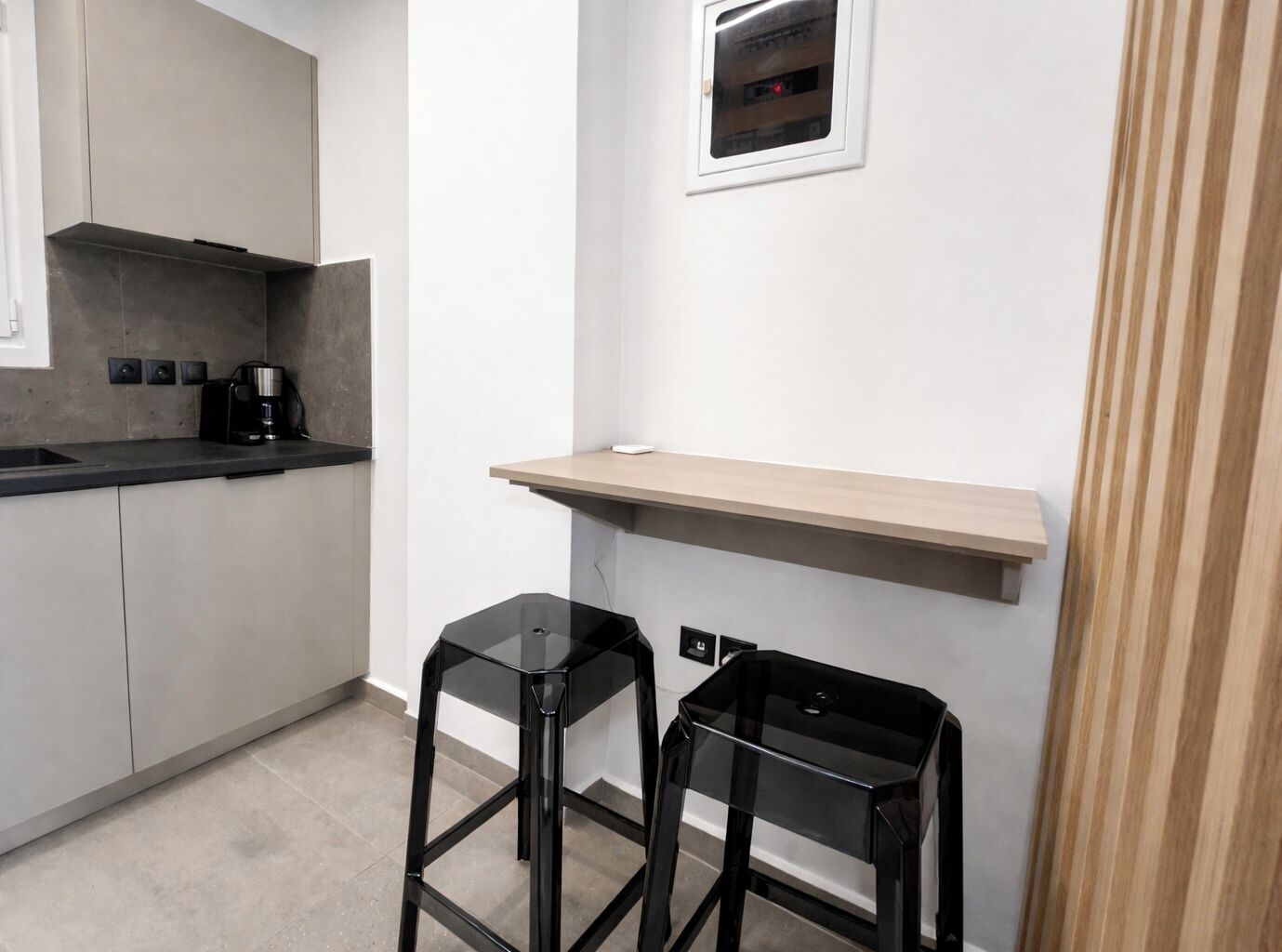 A modern kitchen with a built-in oven, a coffee machine, and a wooden shelf. There are two black stools and a small window with a view of the outside.