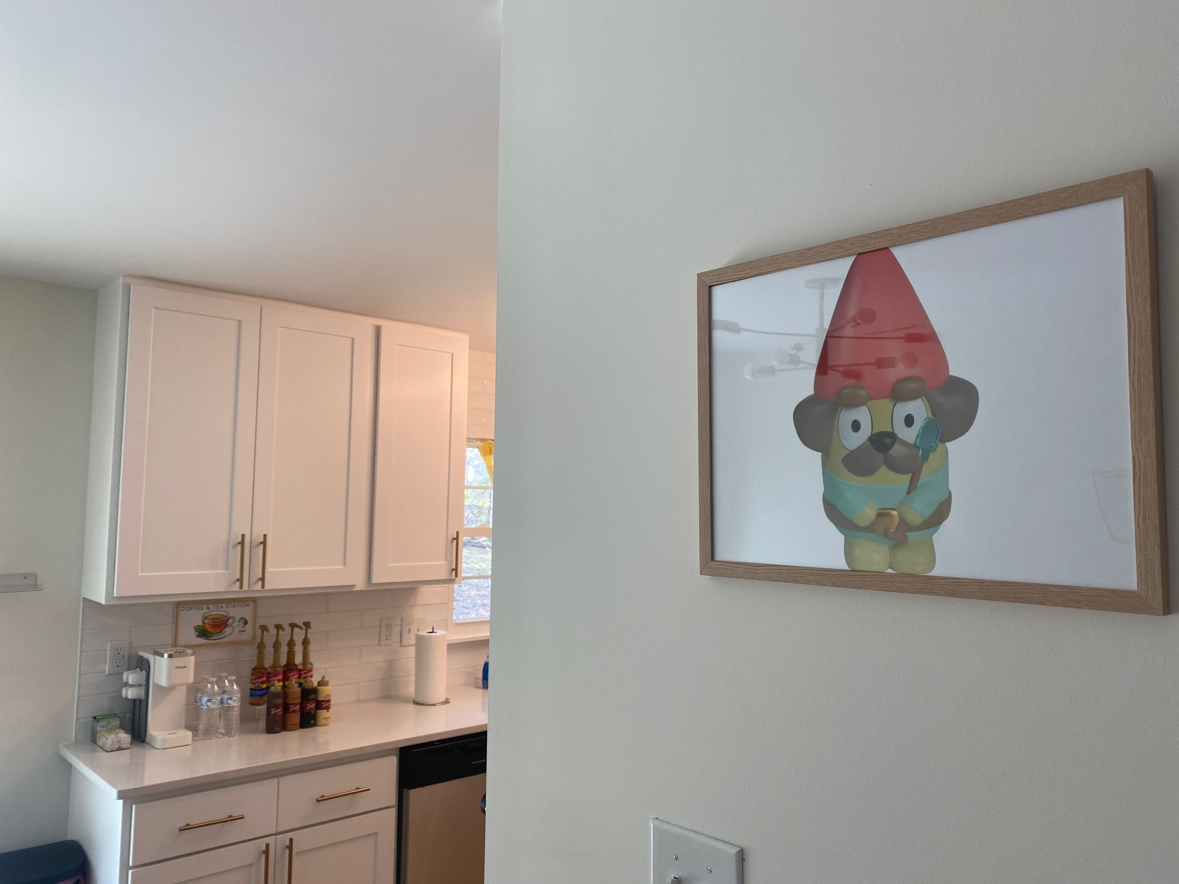 A kitchen with white cabinets, a coffee maker, and a framed cartoon character on the wall.