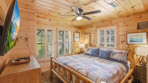 Une chambre dans une cabane en bois, avec un lit, un ventilateur de plafond, une télévision et une fenêtre avec des volets.