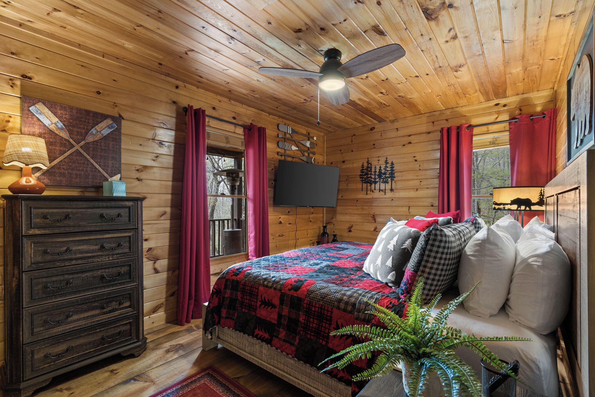 A wooden cabin bedroom with a bed, dresser, TV, and a ceiling fan.