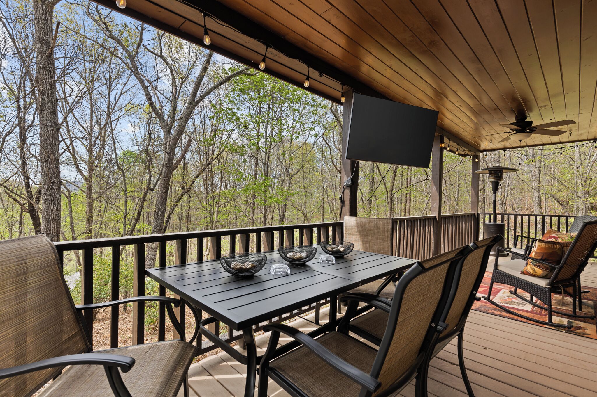 A covered outdoor deck with a table and chairs, a ceiling fan, and a flat-screen TV.