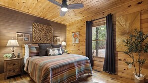 A wooden cabin bedroom with a bed, nightstands, a ceiling fan, and a window with curtains. - Heart's Hideaway - Blue Ridge, GA (Morganton)