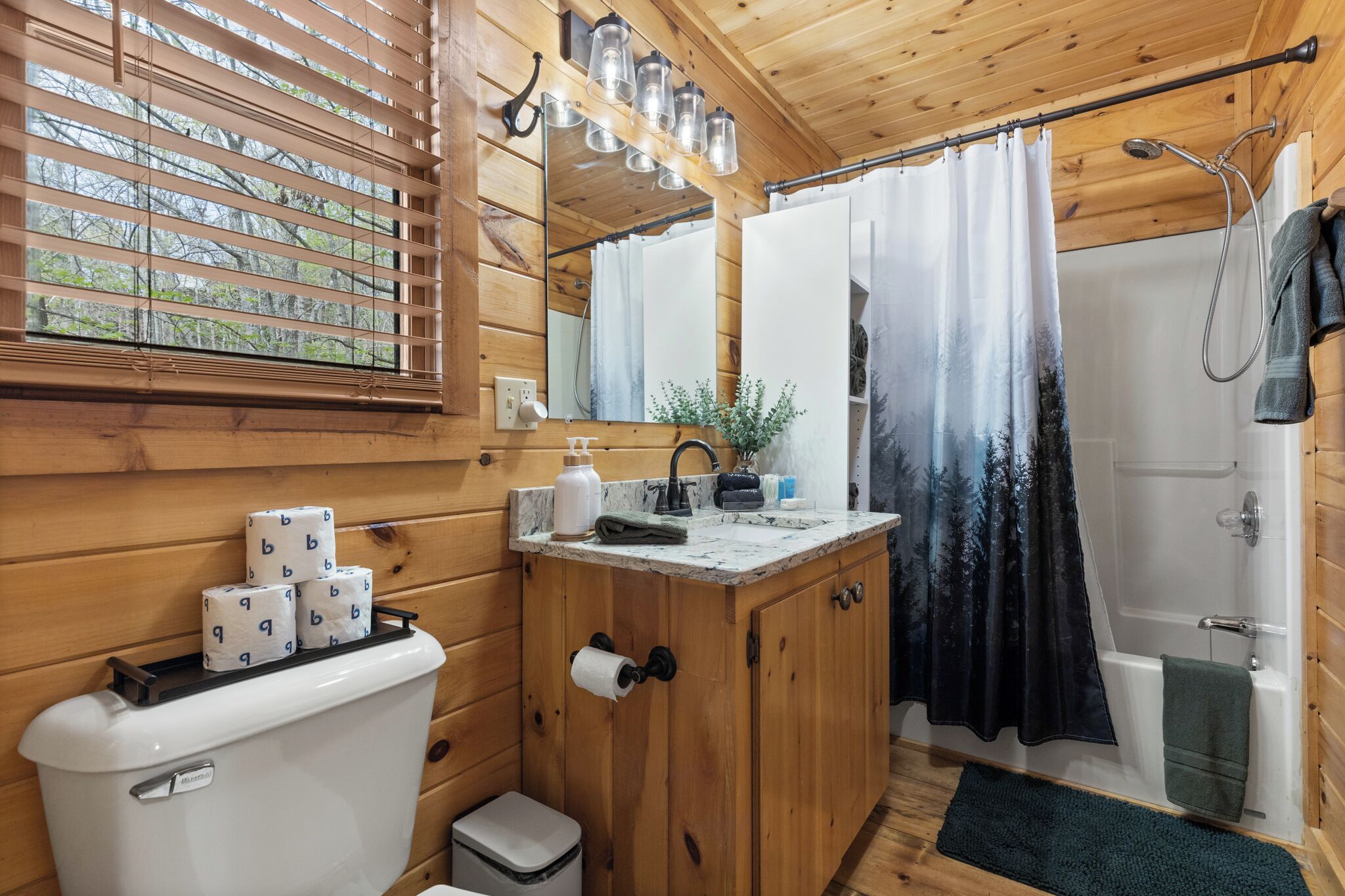 A wooden cabin bathroom with a toilet, sink, shower, and window with blinds.