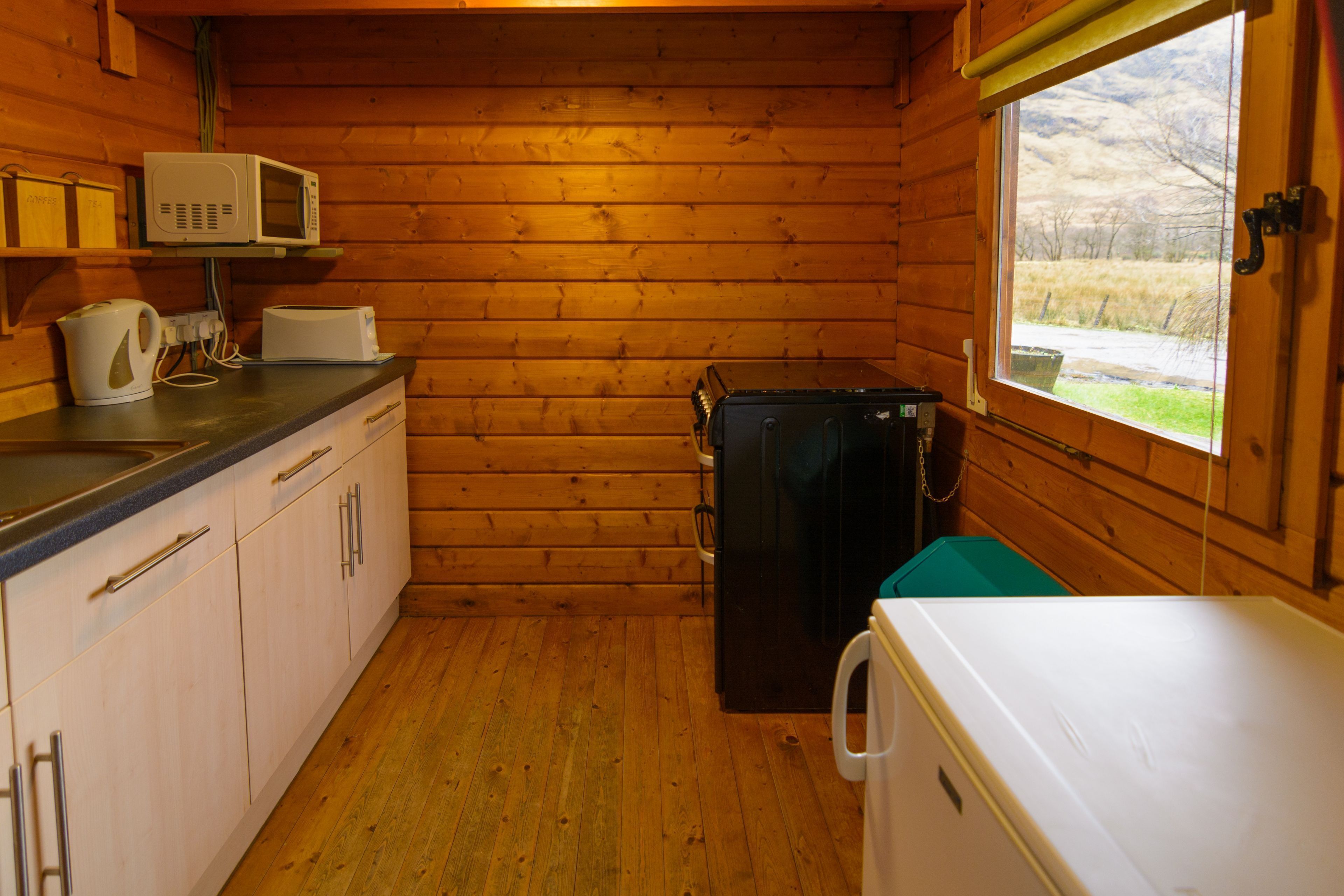Cocina compacta con paredes de madera, microondas, tostadora, refrigerador y ventana con vista al exterior.