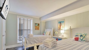 Ein Schlafzimmer mit zwei Betten, einem Fenster mit Jalousien, einem Gemälde, einer Lampe und einem Kissen mit Strandmotiv.
