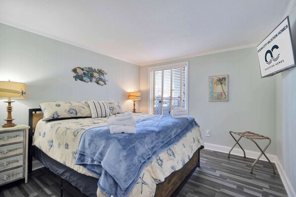 Une chambre à coucher comprenant un lit, deux tables de chevet, une fenêtre avec des stores, un panneau fixé au mur et un petit tabouret.