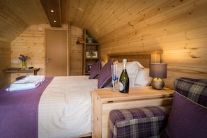 Un dormitorio de cabaña de madera con una cama, una mesita de noche con una botella y vasos, una lámpara y un banco con cojín.