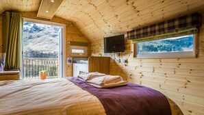 A wooden cabin bedroom with a bed, a window with a view of a mountain, a television mounted on the wall, and a balcony with a potted plant. - Bidean - Cosy Eco Cabin with stunning views (Leacantuim, Glencoe)