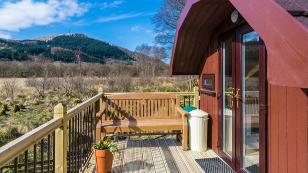 A wooden deck with a bench, a potted plant, and a door to a building with a red exterior. - Bidean - Cosy Eco Cabin with stunning views (Leacantuim, Glencoe)