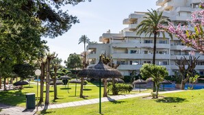 Pátio de hotel com caminho, palmeiras, estrutura de telhado de palha e piscina.