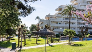 A hotel courtyard with a pathway, palm trees, a thatched-roof structure, and a pool. - Rental BB 436-3 in Benalm Dena (Benalmádena)