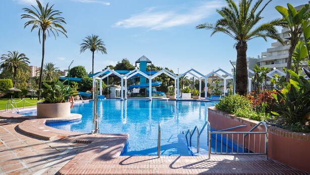 A swimming pool with a blue water slide, palm trees, and a building with a blue roof in the background. - Rental BB 430-3 in Benalm Dena (Benalmádena)