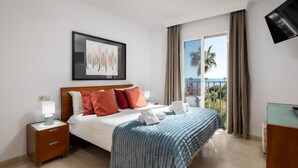 Quarto de hotel com cama, mesa de cabeceira, televisão e varanda com vista para palmeiras e o mar.