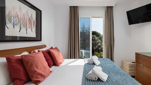 Quarto de hotel com cama, televisão, janela com cortinas e um quadro na parede, com vista para árvores lá fora.